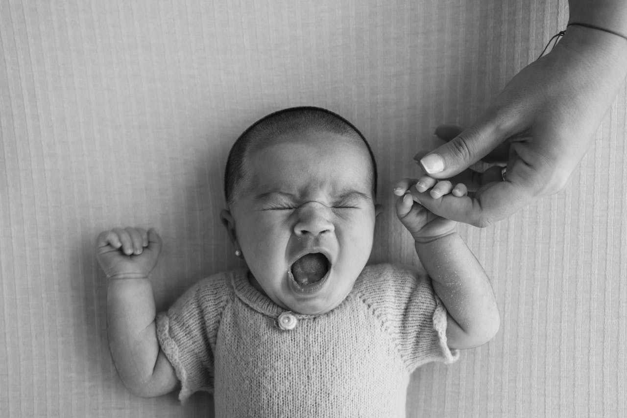 A black and white photo of a yawning newborn holding a hand, conveying warmth and connection.